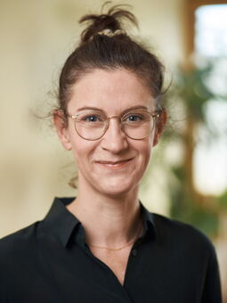 Melanie Wölwer hat das Haar zu einem Dutt zurückgebunden und trägt eine goldene, runde  Metallbrille und ein dunkelgrünes Hemd. Sie lächelt in die Kamera