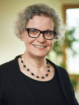 Christiane Rupp hat kurzes, graues, lockiges Haar. Sie trägt ein schwarzes Oberteil und eine Kette mit schwarzen Perlen sowie eine schwarze Brille.