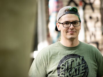 Ein junger Mann  mit Brille und umgedrehter Baseballkappe schaut freundlich in die Kamera. Er trägt ein T-Shirt mit der Aufschrift "Sit ´n Skate"