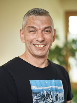 Boris Colić trägt ein schwarzes T-Shirt mit einem Bild einer Stadt darauf. Sein graues Haar ist kurzgeschnitten. Er lächelt in die Kamera