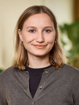 Judith Müller im Porträt. Sie hat blondes, halblanges Haar, das sie mit einem Mittelscheitel trägt. Sie ist gekleidet mit einem braunen T-Shirt und einer etwas helleren, braun melierten Strickjacke.