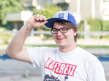 Ein junger Mann mit Baseballkappe lächelt in die Kamera. Seine rechte Hand ist an den Schirm der Mütze gelegt, so als wolle er "den Hut ziehen"