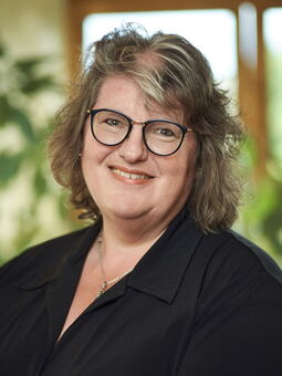 Lydia Wiebalk trägt ein schwarzes Oberteil und eine Silberkette. Sie hat graues Haar mit einer hellen Strähne im Pony und trägt eine schwarze Brille. Sie lächelt in die Kamera