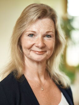 Britta Block hat schulterlanges, blondes Haar mit einem leichten Pony und Sommersprossen im Gesicht. Sie trägt ein weißes Oberteil und einen dunkelblauen Blazer und eine Silberkette. Sie lächelt freundlich in die Kamera.