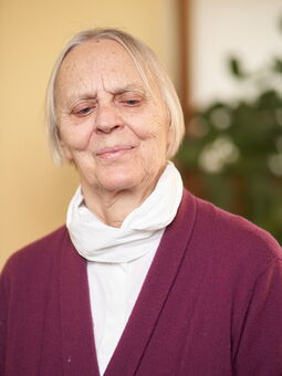 Anke Holtmann trägt ihr weißes Haar zum Bob mit Mittelscheitel. Sie lächelt in die Kamera. Gekleidet ist sie mit einem weißen Rollkragen-Shirt und einer burgunderfarbenen Strickjacke.