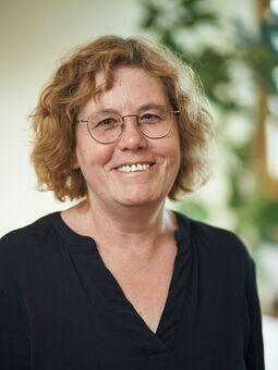 Olga Warnke hat rötlich-blondes, welliges Haar, das sie zu einem Bob mit Seitenscheitel trägt. Dazu eine dünngerahmte runde Brille. Gekleidet ist sie mit einem schwarzen Oberteil mit einem V-Ausschnitt.