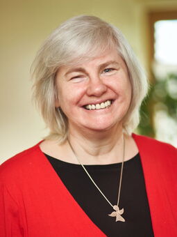 Marion Bonken lächelt strahelnd in die Kamera. Sie hat weißes Haar, dass zum Bob geschnitten ist mit Pony. Auf ihrem schwarzen Oberteil liegt eine Silberkette mit einem Engel als Anhänger. Darüber trägt sich eine rote Jacke.