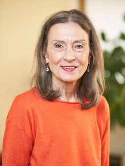 Porträt von Regina Angerstein, die in die Kamera lächelt. Sie hat schulterlanges, braun-graues Haar und trägt einen organgefarbenen Pullover, tropfenförmige Ohrringe und roten Lippenstift.