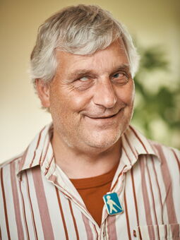 Clemens Gojowczyk hat kurzes graues Haar und lächelt verschmitzt in die Kamera. Gekleidet ist er mit einem rot-weiß gestreiften Hemd über einem roten T-Shirt. Ein Anstecker mit dem Stockgänger ist an seinem Rever befestigt.