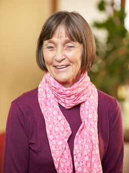 Margarete Schulze trägt einen burgunderfarbenen Pullover und ein rosafarbenes Halstuch mit weißen Blüten darauf. Ihr braun-graues Haar ist zu einem Bob geschnitten, sie trägt einen Pony.