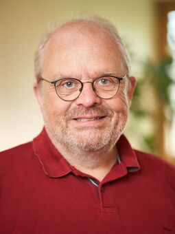 Achim Becker im Porträt. Er trägt einen roten Pullover mit Polokragen und eine schwarzrandige Brille. Sein Haar ist weiß, mit einem hohen Ansatz und er hat einen Dreitagebart.