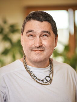 André Rabe im Porträt. Er trägt ein weißes T-Shirt und große Ketten. Er schaut freundlich in die Kamera.