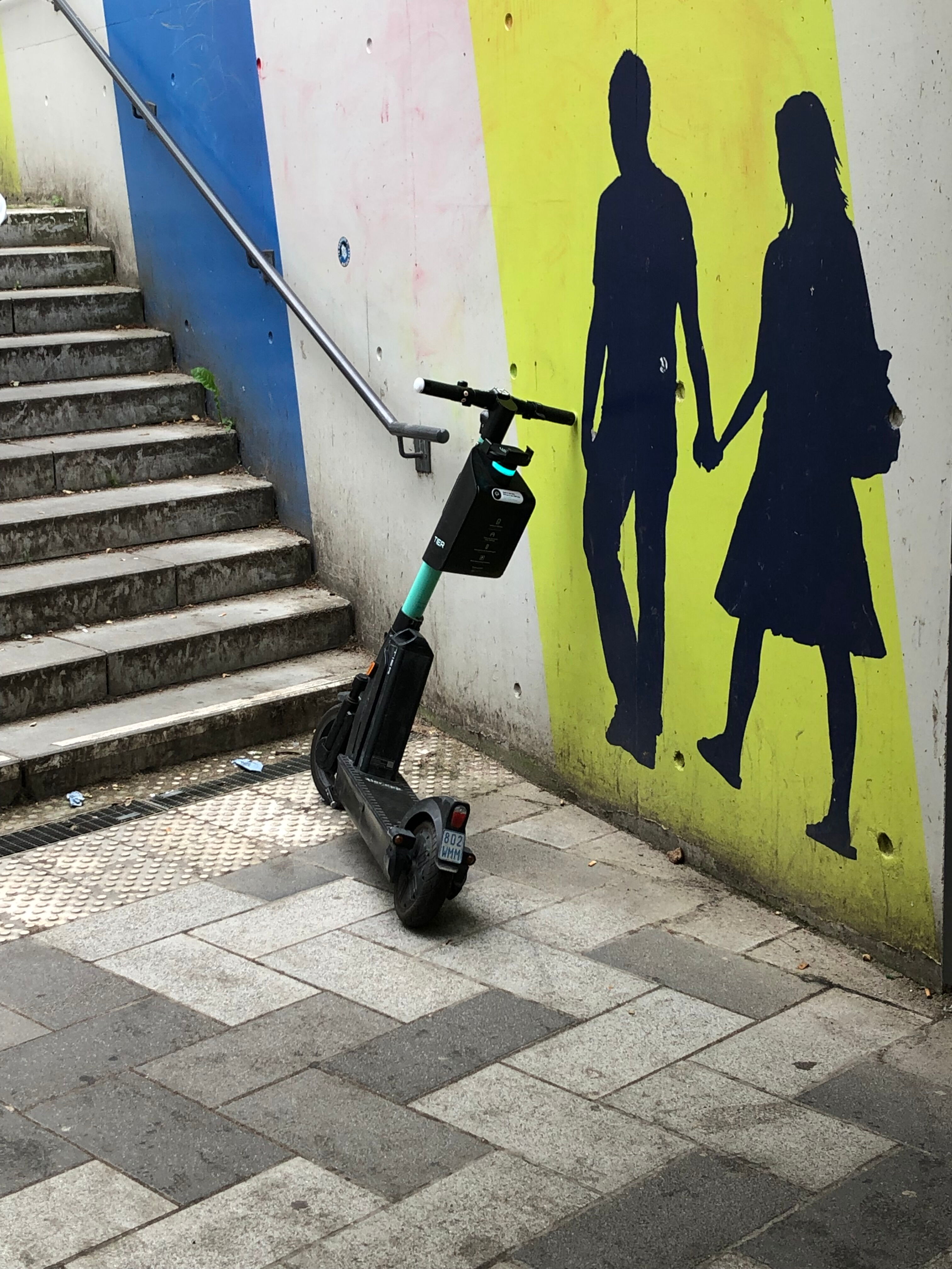 Ein E-Roller in einer Bahnstation vor dem Treppenaufgang. Er versperrt den Weg zum Handlauf an der Treppe