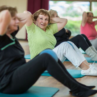 Mehrere ältere Frauen machen Turnübungen nebeneinander auf Matten in einer Turnhalle