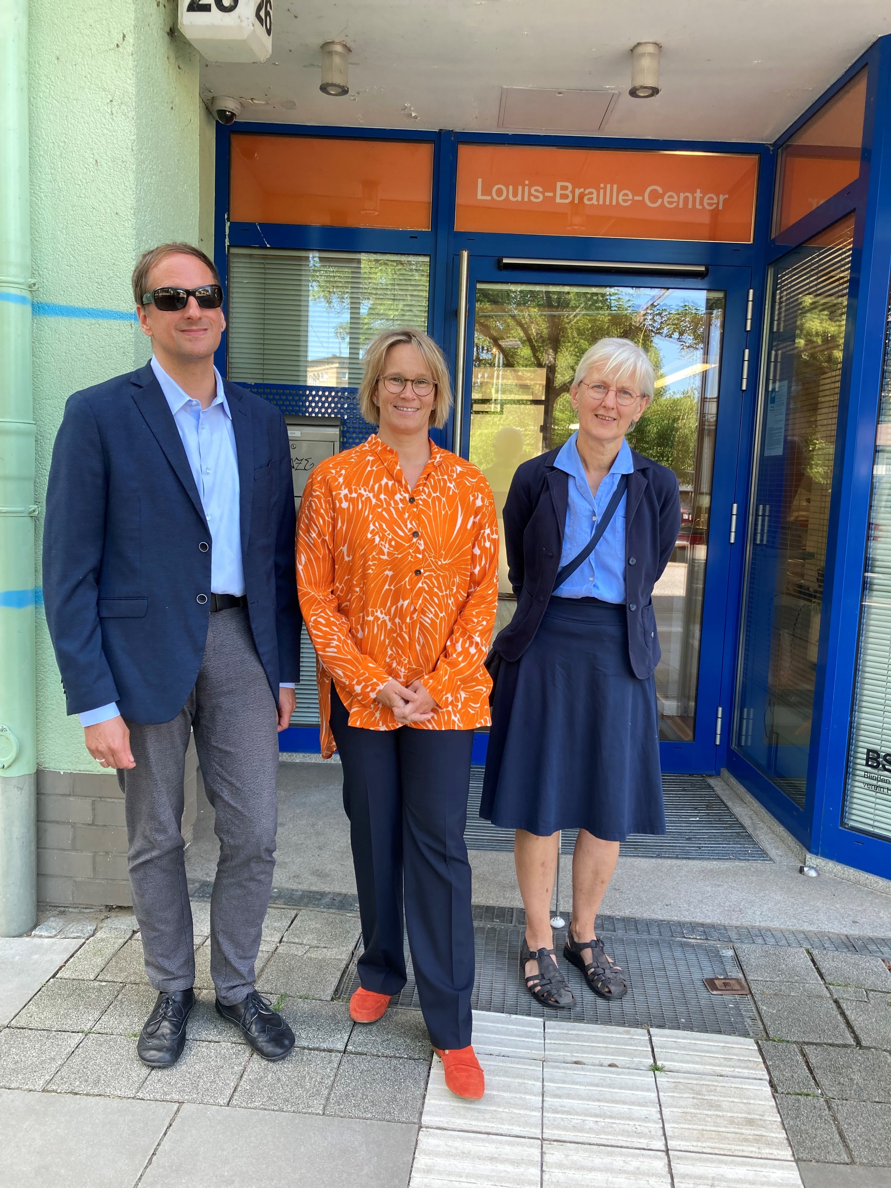 Die Sozialsenatorin Melanie Schlotzhauer steht zwischen BSVH Geschäftsführer Heiko Kunert (li.) und der 1. Vorsitzenden des BSVH, Angelika Antefuhr (re.) vor dem Eingang des Louis-Braille-Centers