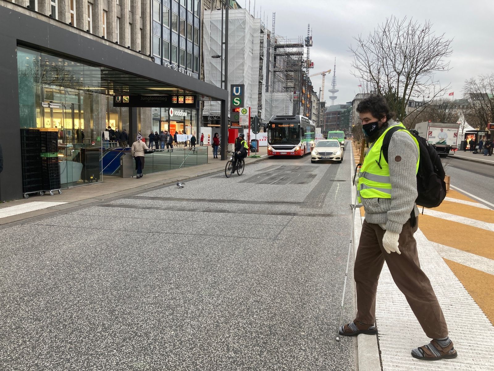 Ein blinder Mann tritt an einer ungesicherten Querung auf die Straße am umgebauten Jungfernstieg. Im Hintergrund nähern sich ein Bus, ein Taxi sowie ein Fahrrad.
